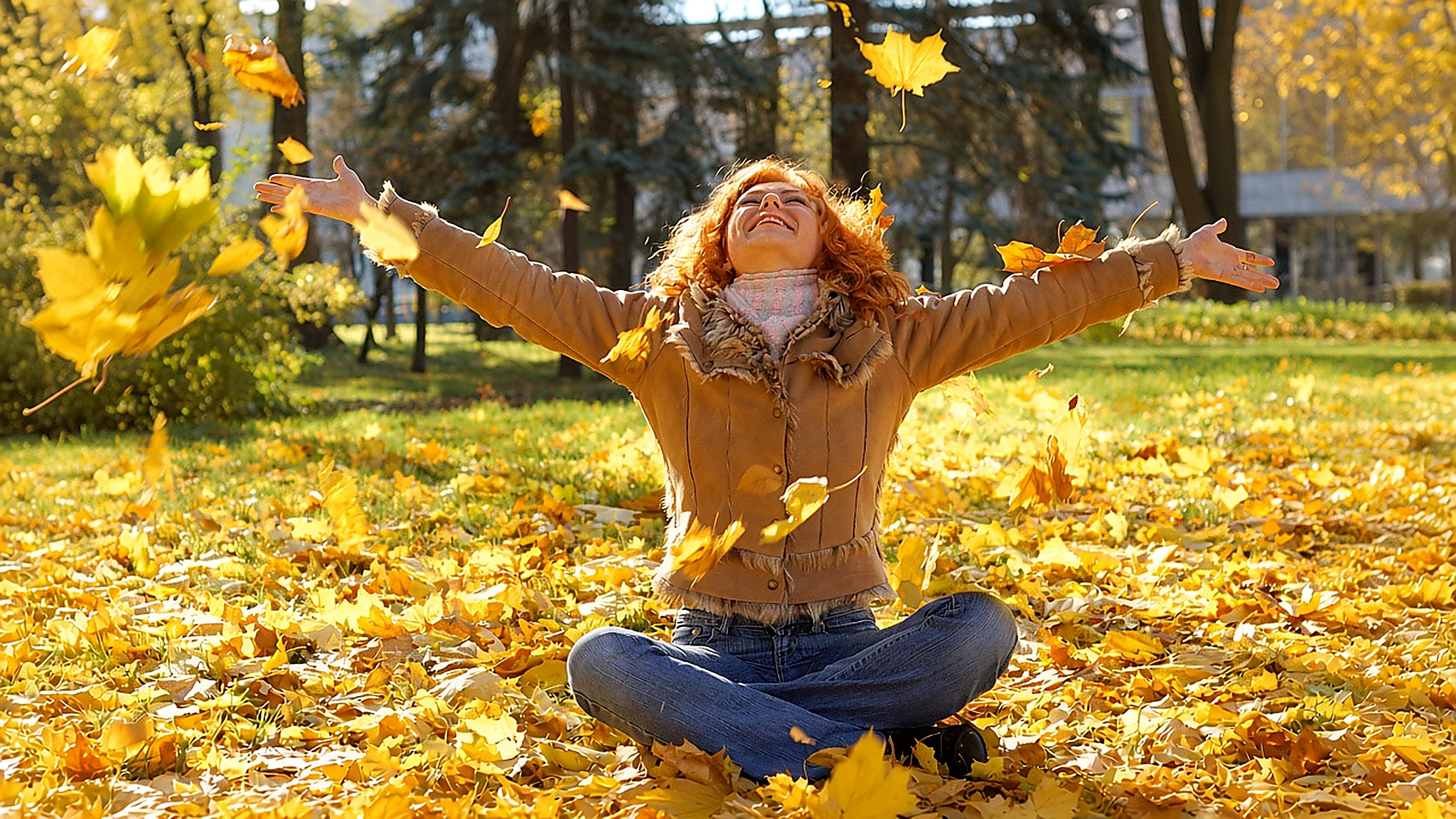 Donna sorridente seduta tra le foglie gialle in un parco autunnale, con giacca color cammello e jeans