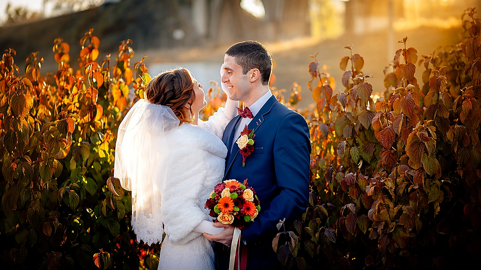 Sposi abbracciati tra foglie rosse autunnali, lei con stola bianca e bouquet arancio, lui in abito blu
